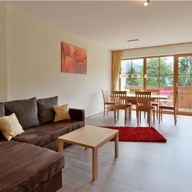 Ein gemütliches Wohnzimmer mit einer Couch, einem Tisch und Stühlen. Große Fenster bieten viel Tageslicht und einen Blick nach draußen.