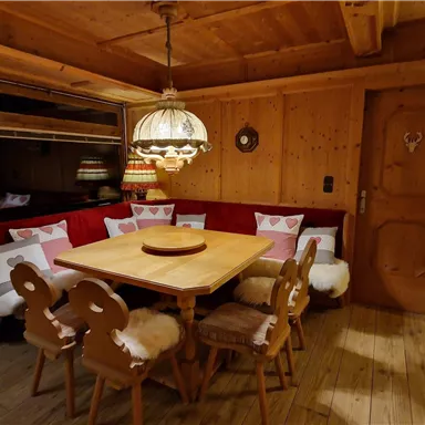 Eine gemütliche Stube mit Holzverkleidung und einem runden Tisch in der Mitte. Um den Tisch stehen Stühle mit Fellbezügen und eine einladende Bank mit Kissen.