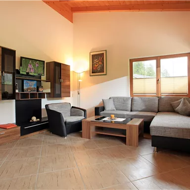 Ein modernes Wohnzimmer mit bequemen Möbeln und großen Fenstern. Eine gemütliche Atmosphäre wird durch die Holzverkleidung und die dekorativen Elemente geschaffen.