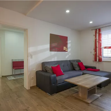 A modern living room with a gray sofa and red pillows. The room has a warm wooden floor and large windows with curtains.