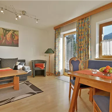 Ein gemütlicher Wohnraum mit einem grauen Sofa und einem Holztisch. Der Raum ist hell und freundlich gestaltet, mit bunten Vorhängen und einem Blick auf die Außenansicht.