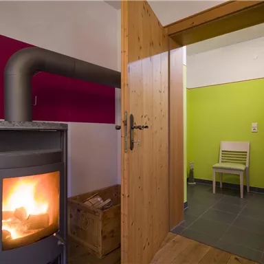 A cozy room with a wood stove and colorful walls. In the background, a chair is visible on a tiled floor.