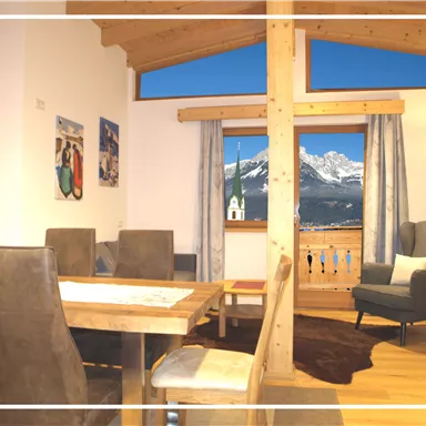 Ein gemütlicher Wohnraum mit Holzdecken und einem großen Fenster, das einen Blick auf die Berge bietet. Der Raum ist einfach eingerichtet mit einem Tisch, Stühlen und einer bequemen Ecke.