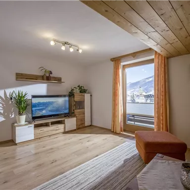 A cozy living room with a wooden ceiling and large windows. There is a modern TV corner and plants that create an inviting atmosphere.