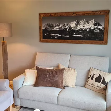 A cozy living room with a gray sofa and various cushions. A large landscape painting of mountains hangs on the wall.