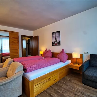 Ein helles Zimmer mit zwei Betten, einem blauen Sofa und bequemen Sesseln. Die Wände sind neutral, und es gibt ein Bild von Paris.