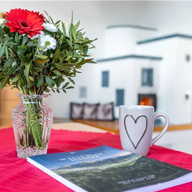 A laid table with a vase full of flowers and a cup of coffee. In the background lies a book with a nature landscape on a red table runner.
