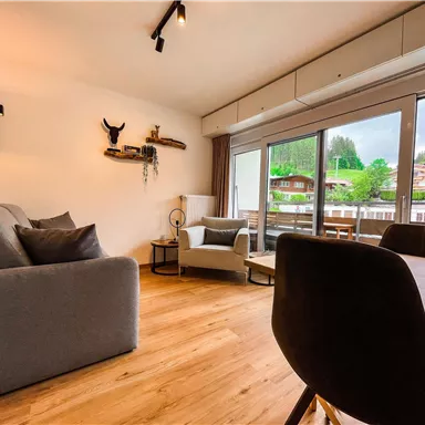 Ein modernes Wohnzimmer mit einem grauen Sofa und einem Esstisch. Große Fenster bieten einen Blick ins Grüne.