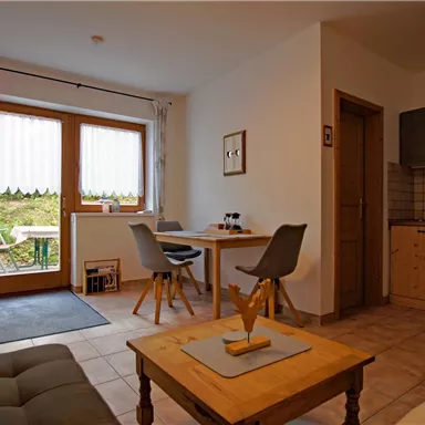 A cozy living room with bright walls and large windows. There is a dining table, a couch, and a modern kitchen.