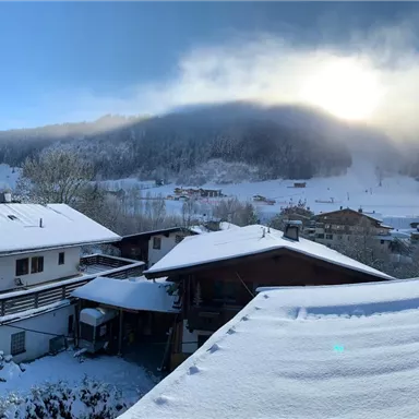 A picturesque winter scene with snow-covered roofs and mountains in the background. The sun shines through the clouds, giving the landscape a warm light.