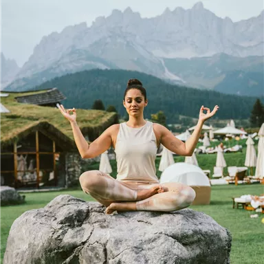A woman meditates peacefully on a large rock. In the background, mountains and a green landscape can be seen.