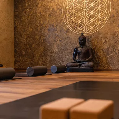 Ein ruhiger Yogaraum mit einer sitzenden Buddha-Statue und einer dekorativen Wand. Matten und Yogazubehör liegen bereit für eine entspannende Sitzung.