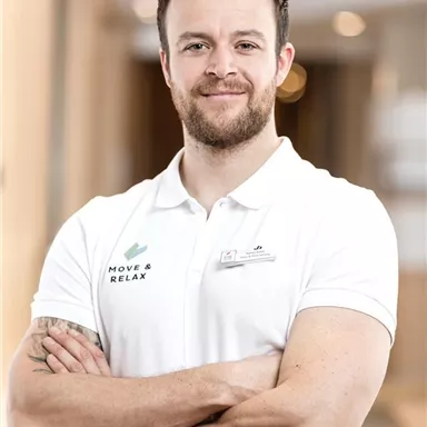 Ein sportlich aussehender Mann in einem weißen Poloshirt steht mit verschränkten Armen. Er lächelt freundlich und wirkt einladend.