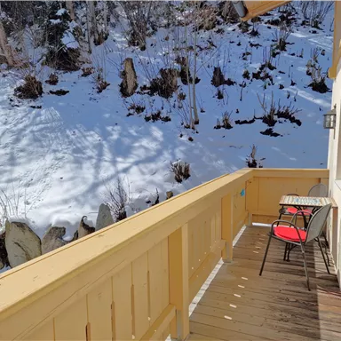Ein Holzbalcony mit einem Tisch und zwei Stühlen blickt auf eine schneebedeckte Landschaft. Die Sonne scheint auf die verschneite Fläche im Hintergrund.