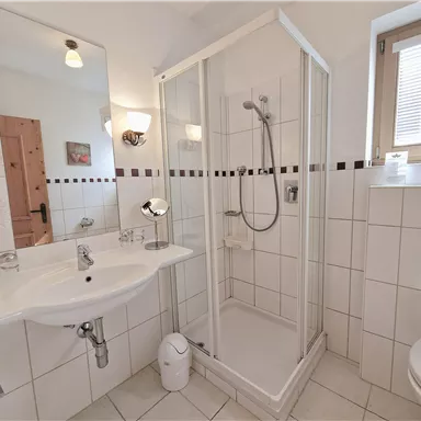 Ein modernes Badezimmer mit einer Dusche, einem Waschbecken und einem WC. Die Wände sind mit weißen Fliesen gefliest und es gibt viel Tageslicht.