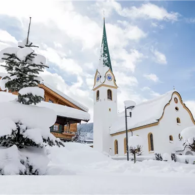 A snowy landscape with a beautiful white church and a green tree. The sky is clear and there are a few clouds.