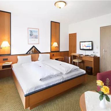A simple hotel room with a double bed, a television, and a small table. There is also a red armchair and windows that let in a lot of light.