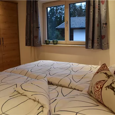 Ein gemütliches Schlafzimmer mit einem großen Fenster und einem Holzkleiderschrank. Das Bett ist mit weißen, gemusterten Decken und dekorativen Kissen ausgestattet.