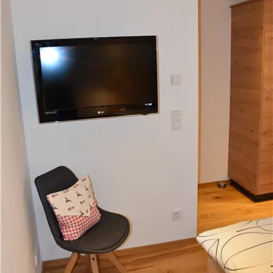 Ein gemütlicher Raum mit einem Fernseher an der Wand und einem Stuhl mit einem Kissen. Der Boden ist aus Holz und die Möbel sind modern.