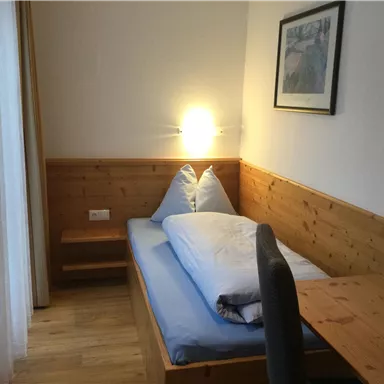 Ein gemütliches Zimmer mit einem Einzelbett, einem Schreibtisch und schönen Holzdetails. Die Wände sind hell und es gibt ein Fenster mit Vorhängen.