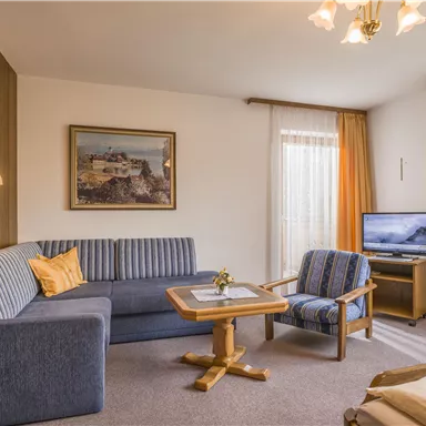 A cozy living room with a blue couch and a small table. Large windows with curtains bring a lot of light into the room.