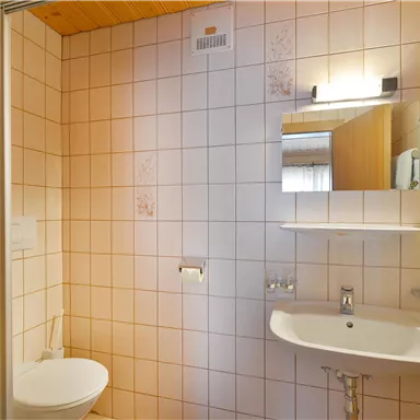 A small bathroom with tiled walls and a sink. There is a toilet and a mirror above the sink.