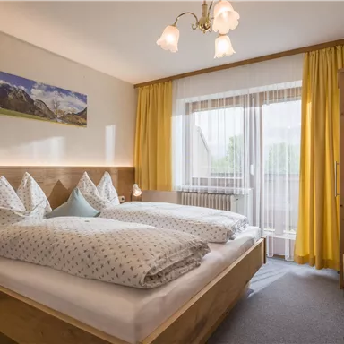 A cozy bedroom with two beds and large pillows. Bright windows and yellow curtains create a friendly atmosphere.