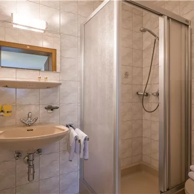 A simple bathroom with a shower, sink, and mirror. The walls are tiled, and the furnishings are functional.