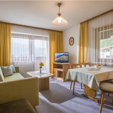 A cozy living room with a sofa, a table, and chairs. Large windows let in plenty of light and offer a view of the surroundings.