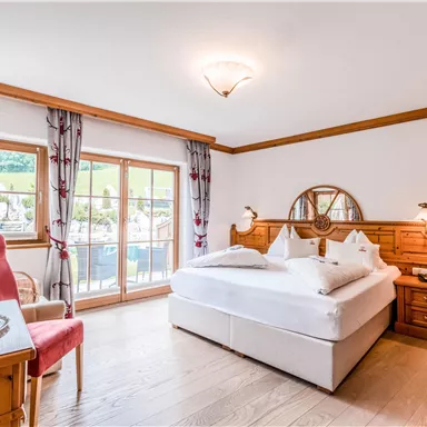 Ein gemütliches Schlafzimmer mit einem großen Bett und warmen Holzmöbeln. Große Fenster lassen viel Tageslicht herein und bieten einen Blick ins Freie.