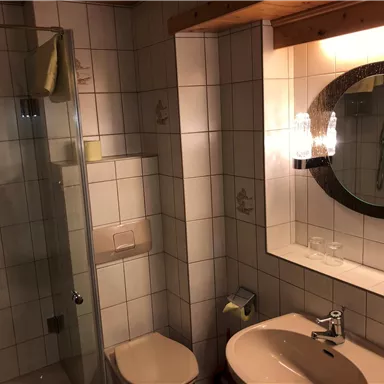 Ein einfaches Badezimmer mit Dusche, Waschbecken und Spiegel. Die Wände sind gefliest und die Ausstattung ist funktional.