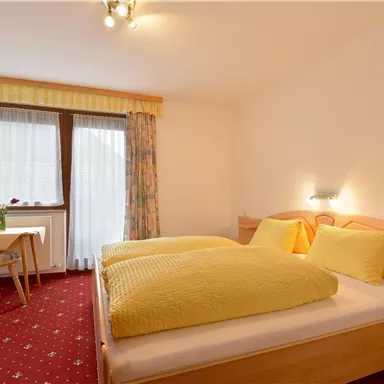 A cozy bedroom with two beds and yellow pillows. In the corner stands a table with chairs and large windows that provide plenty of light.
