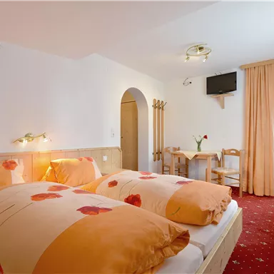 A cozy bedroom with two beds and decorative pillows. The room has a television and a small dining table, as well as light curtains.