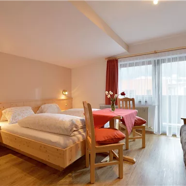 A simple room with two beds, a dining table, and a sofa bed. Large windows provide plenty of daylight and a pleasant atmosphere.