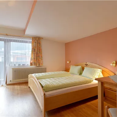 A cozy bedroom with two beds and a table. The room has large windows that provide natural light.