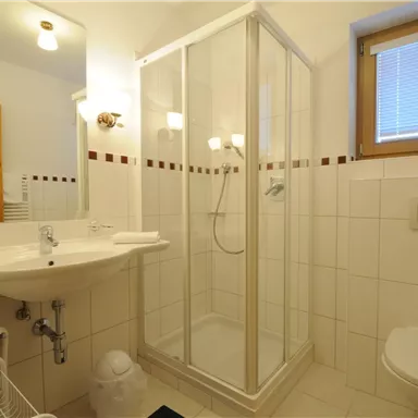 Ein helles Badezimmer mit einer Glasdusche und einem Waschbecken. Die Wände sind gefliest und es gibt ein Fenster für Tageslicht.