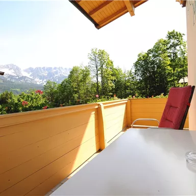 Ein Balkon mit einem Tisch und einem Stuhl. Im Hintergrund sind Bäume und Berge zu sehen.