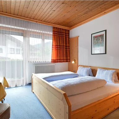 Ein gemütliches Zimmer mit einem Doppelbett und Holzdekor. Große Fenster sorgen für viel Tageslicht und eine angenehme Atmosphäre.