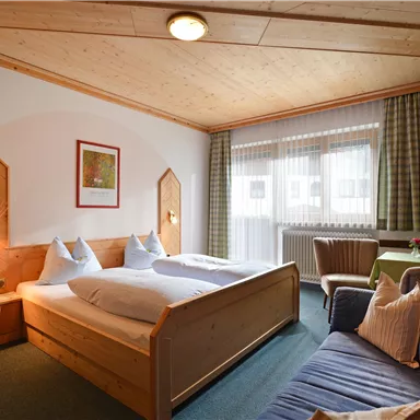 Ein gemütliches Zimmer mit zwei Betten und einem Holzdekor. Es gibt einen bequemen Sessel und viel Tageslicht durch die Fenster.