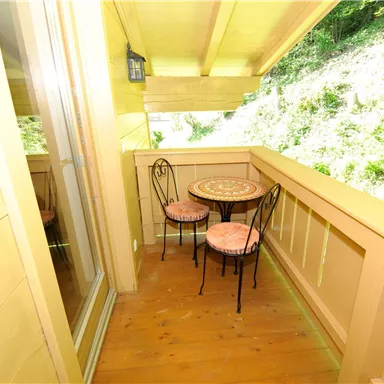 Ein kleiner Balkon mit einem Tisch und zwei Stühlen. Die Umgebung besteht aus üppigem Grün.