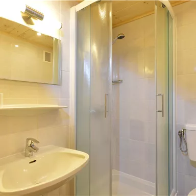 Ein modernes Badezimmer mit einer Dusche, einem Waschbecken und einem Spiegel. Die Wände sind hell gefliest und der Raum wirkt gut beleuchtet.