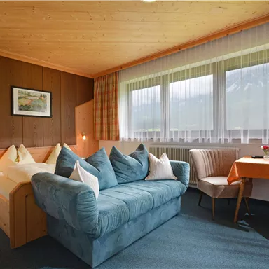 Ein gemütliches Zimmer mit Holzverkleidung und großen Fenstern. Es gibt ein bequemes Sofa, Kissen und einen kleinen Tisch mit Stühlen.