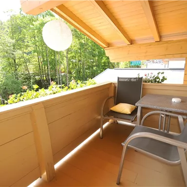Ein gemütlicher Balkon mit einem Tisch und zwei Stühlen. Der Blick geht auf eine grüne, bewaldete Umgebung.