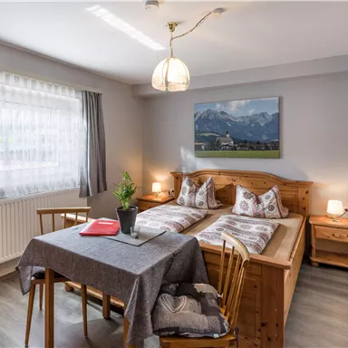 A cozy bedroom with two single beds and a small table. The room has a large window and a decorative wall with a landscape picture.