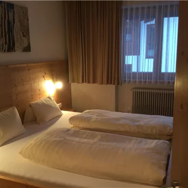 Ein gemütliches Schlafzimmer mit einem Doppelbett und zwei Kissen. Das warm beleuchtete Zimmer hat Holzdetails und große Fenster mit Vorhängen.