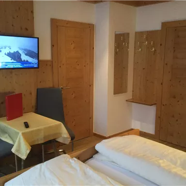 Ein gemütliches Zimmer mit zwei Betten, einem Tisch und Stühlen. Es gibt einen Fernseher an der Wand und eine helle, einladende Atmosphäre.