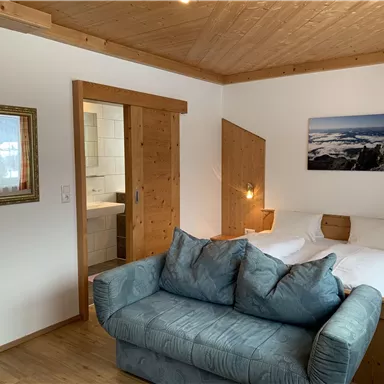 Ein gemütliches Zimmer mit einem Doppelbett und einer blauen Couch. An der Wand hängt ein Bild einer Landschaft, und das Zimmer hat Holzelemente für eine warme Atmosphäre.