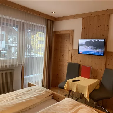 Ein gemütliches Hotelzimmer mit zwei Betten und einem Fernseher an der Wand. An einem kleinen Tisch stehen zwei Stühle und es gibt viel Tageslicht durch die Fenster.