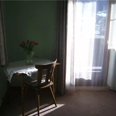 A bright room with a table and a chair. On the table, there is a vase with red tulips and the sun is shining through the windows.