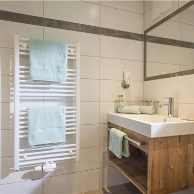 A modern bathroom with a white sink and a wooden cabinet. Two green towels hang on the wall and a towel warmer is available.